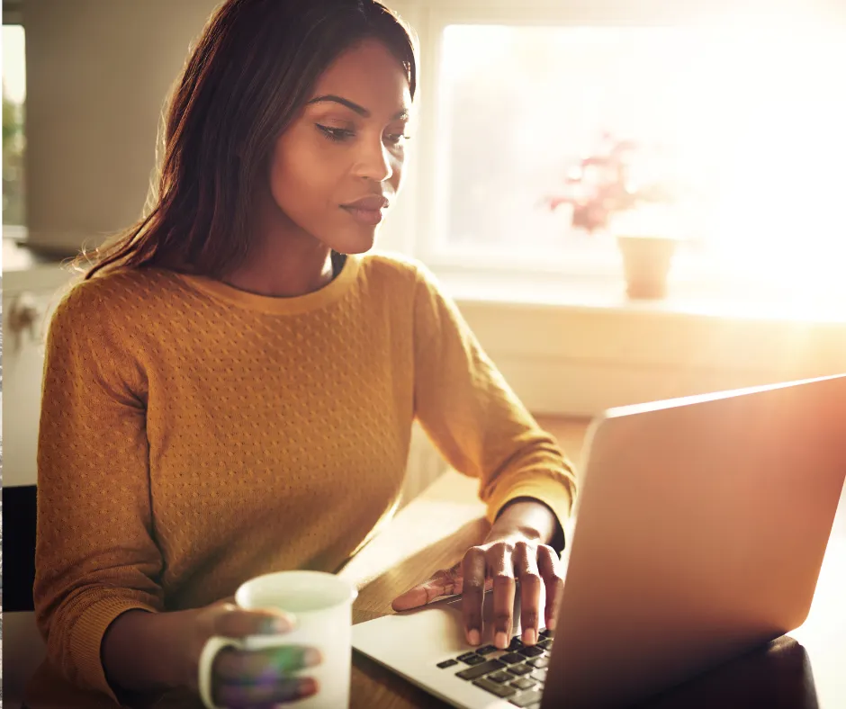 Woman in mustard yellow sweater browsing aesthetic services on laptop at home with coffee, natural window light in background